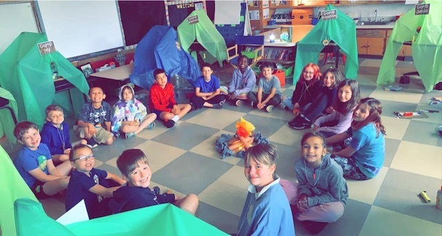 Students sitting by their make-shift tents and fake campfire in their classroom